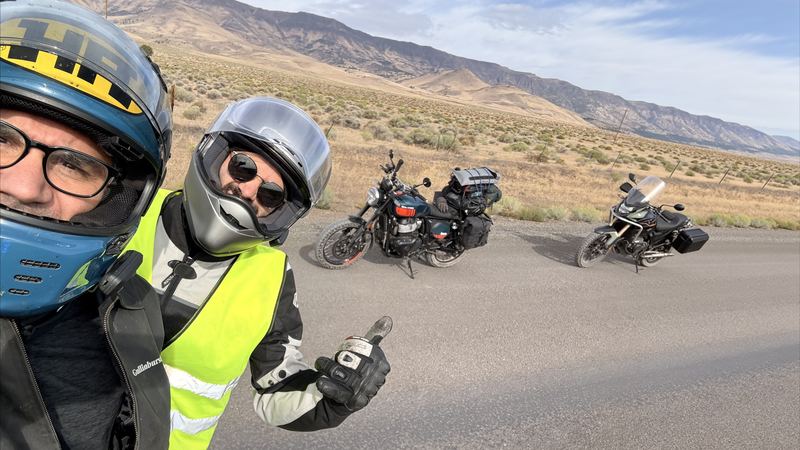 Two riders in helmets taking a selfie on a desert road, their bikes parked behind