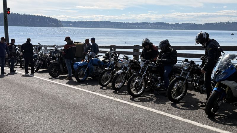 A row of vintage motorcycles, Nortons among them, lined up along a waterfront railing