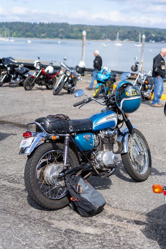 A blue and silver vintage motorcycle parked by the water, with sailboats in the background