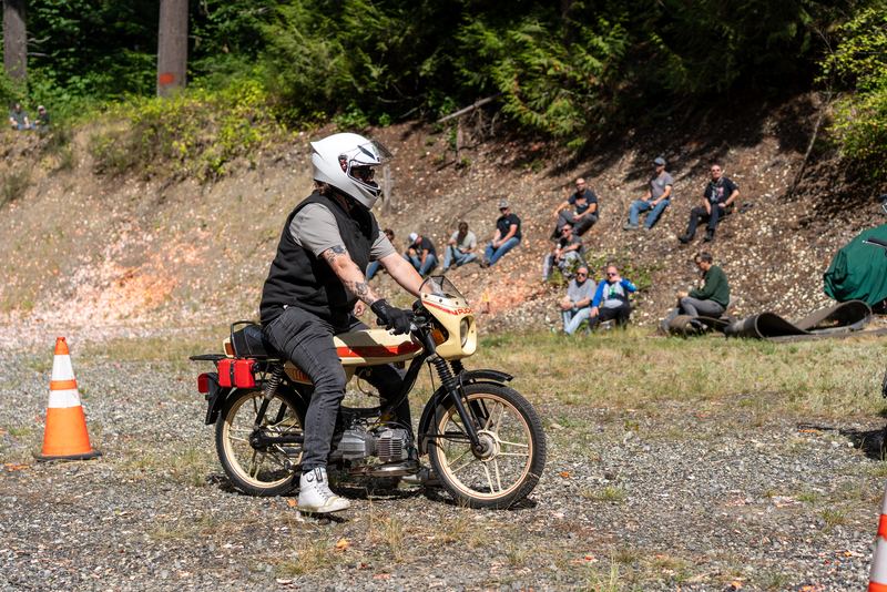 A rider in a white helmet on a small vintage motorcycle on the gravel course, spectators on the hill behind