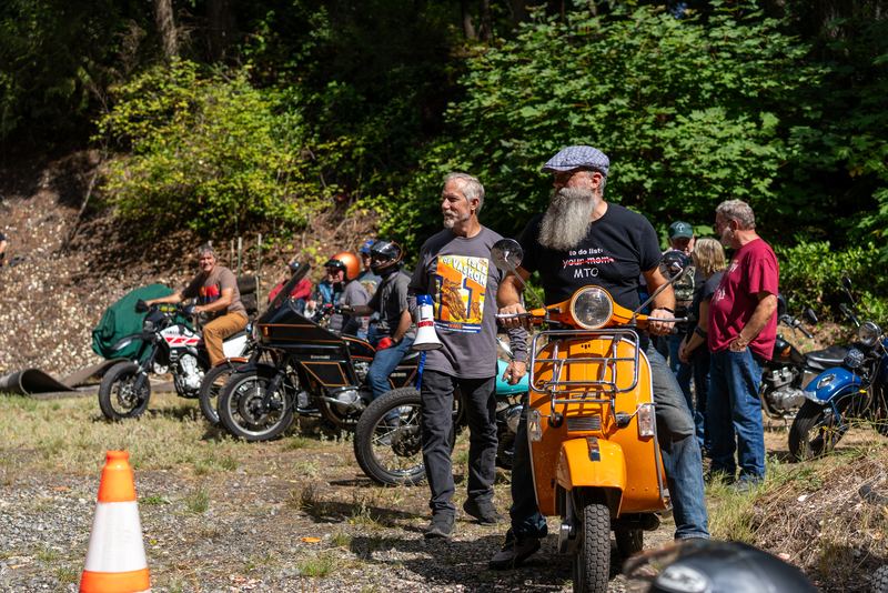 Riders and spectators standing around an orange Vespa scooter on the course