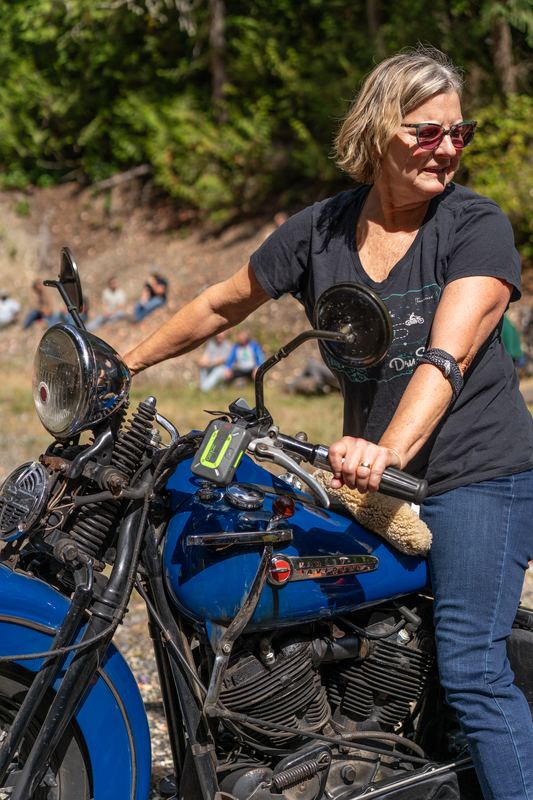 A woman in sunglasses standing over a blue vintage motorcycle, spectators on the hillside behind